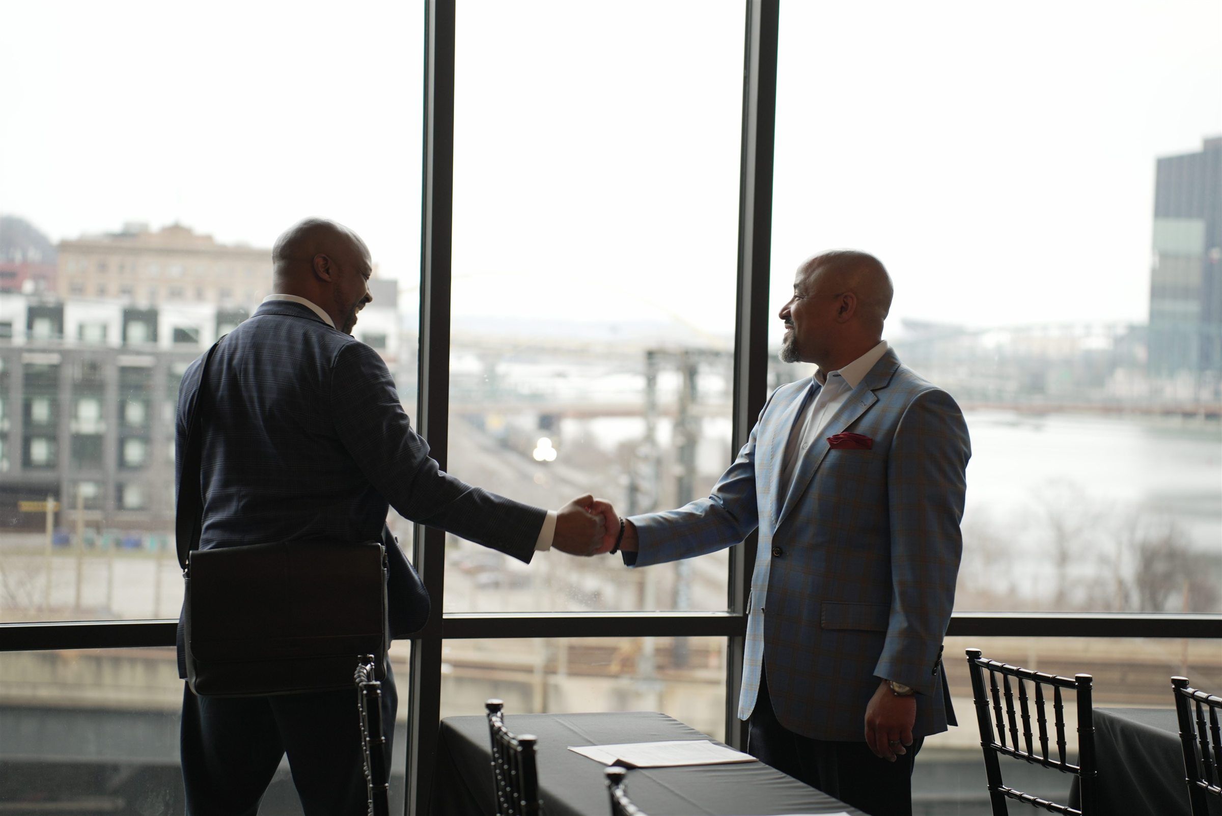 Two business leaders shaking hands in front of windows overlooking the river