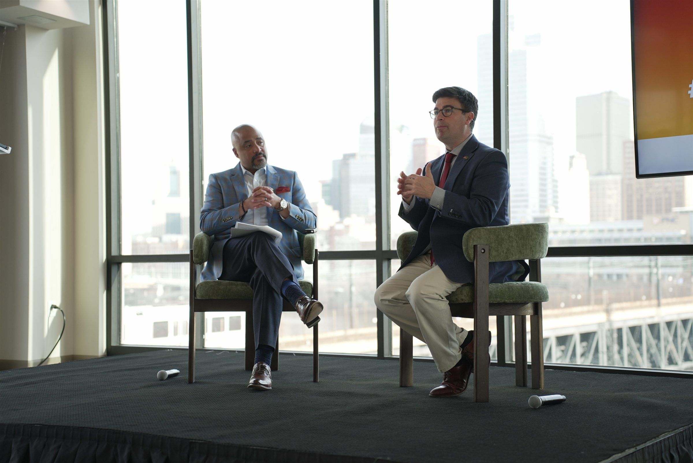 Two speakers in conversation on a panel stage with the Pittsburgh skyline behind them